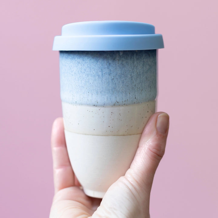 Hand holding a two-toned ceramic cup with a blue lid against a pink background