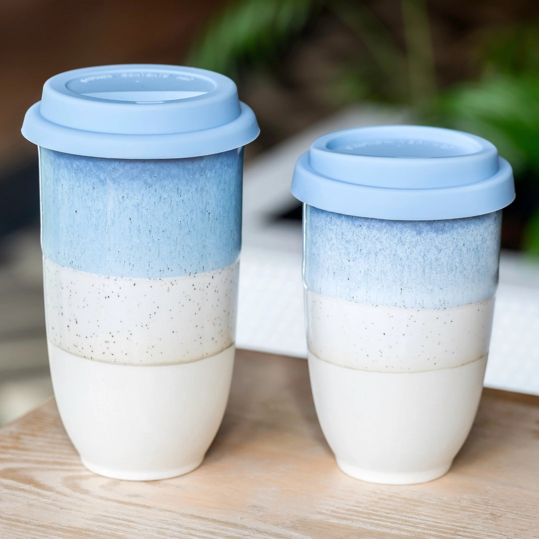 Two blue and white ceramic travel mugs on a wooden surface with a blurred natural background.