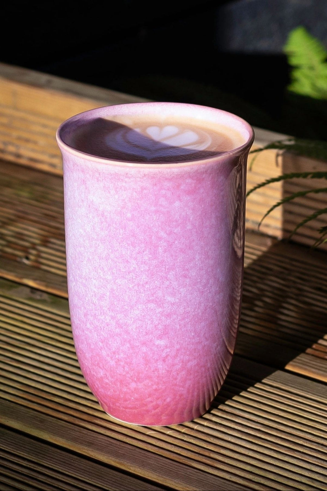 A pink ceramic mug filled with a latte, placed on a wooden slatted surface in bright sunlight.
