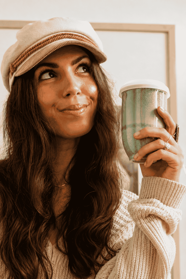 A woman with long wavy brown hair wearing a cream-colored knit sweater and matching cap holds a green glittery insulated coffee cup. She is looking up and smiling in a warm, cozy indoor setting.