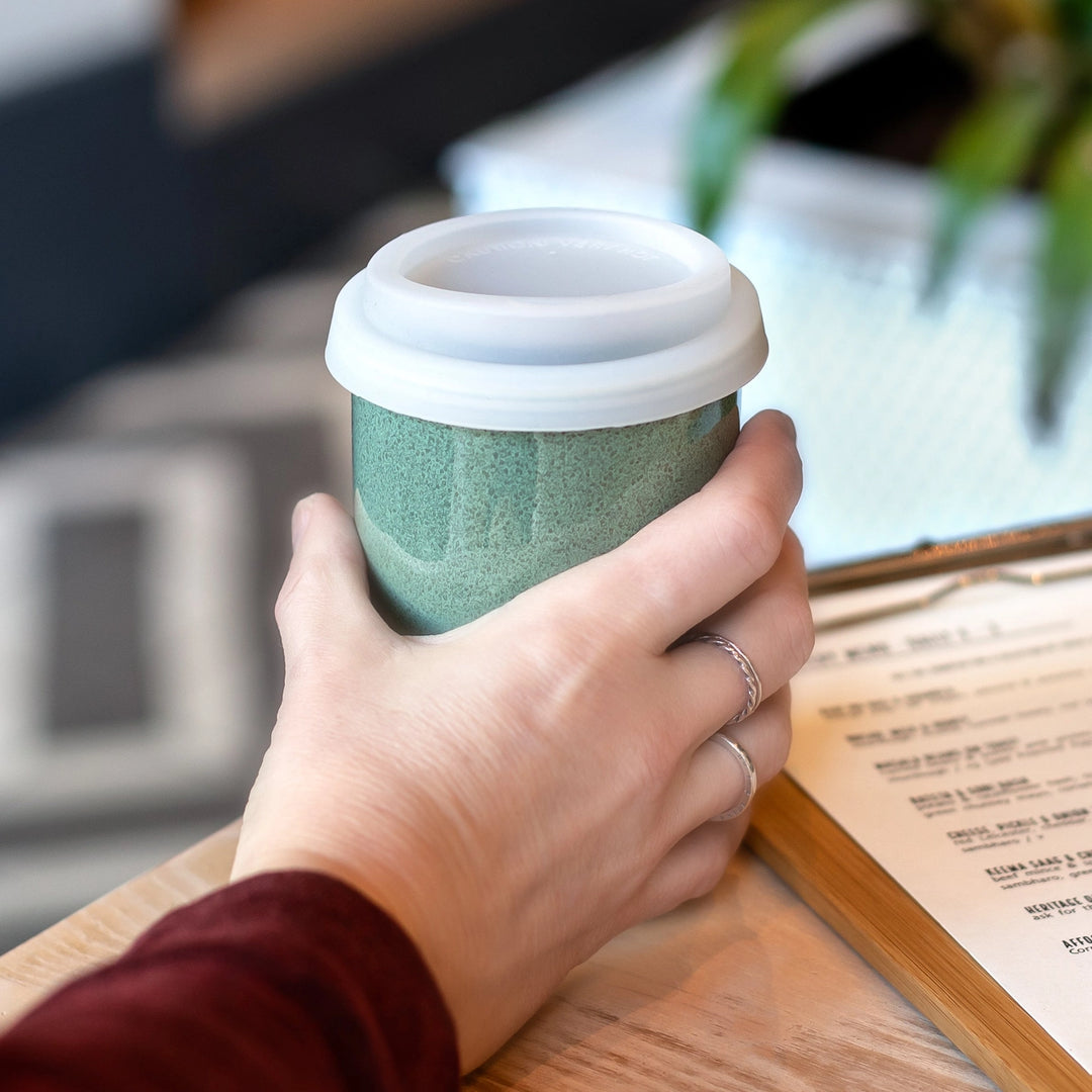 Hand holding a green reusable coffee cup with a white lid on a wooden table.