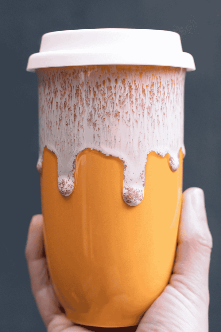 A hand holding a ceramic travel mug with a white lid. The mug features a bright orange lower half and a white upper half with a dripping wax effect, filled with a light-colored beverage.