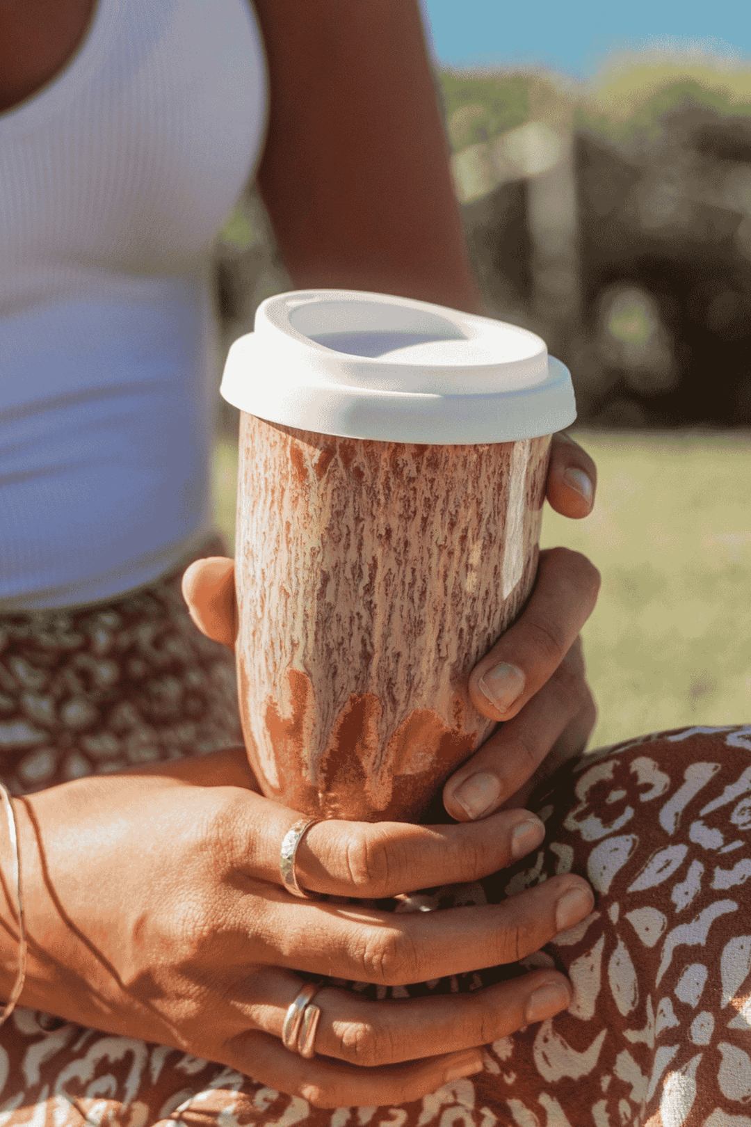 A close-up of a person holding a reusable coffee mug outdoors. The mug has a textured brown and beige bark-like pattern with a white lid.