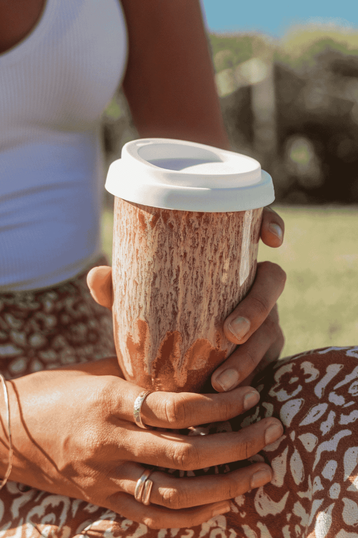 A close-up of a person holding a reusable coffee mug outdoors. The mug has a textured brown and beige bark-like pattern with a white lid.
