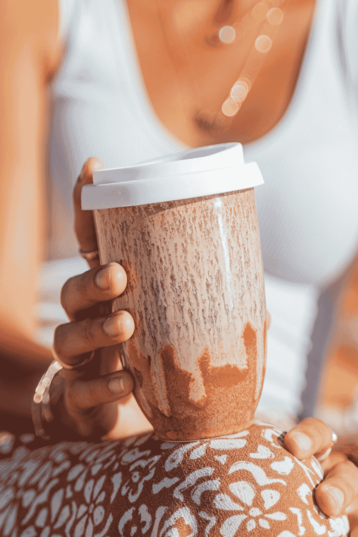 A close-up lifestyle shot of a person holding a brown ceramic travel mug with a white lid, wearing a white tank top and a patterned brown and white skirt.