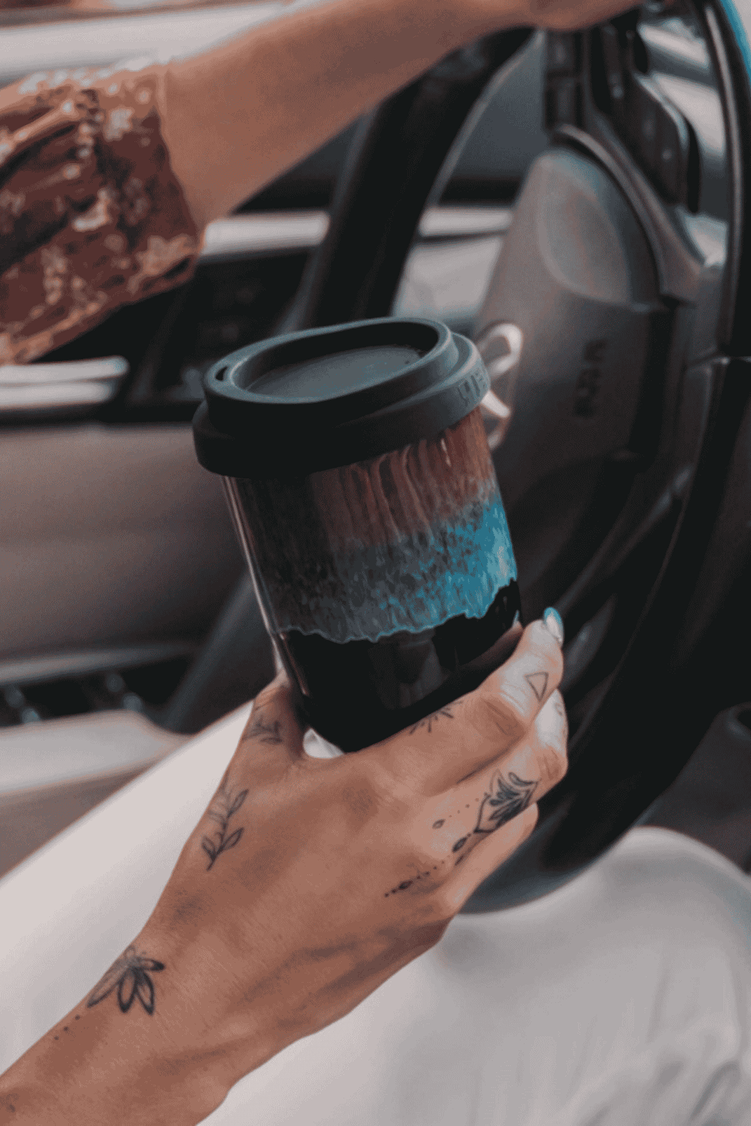 A person sitting in a car holds a reusable coffee cup with a black lid and a dark-to-blue gradient design. The person&