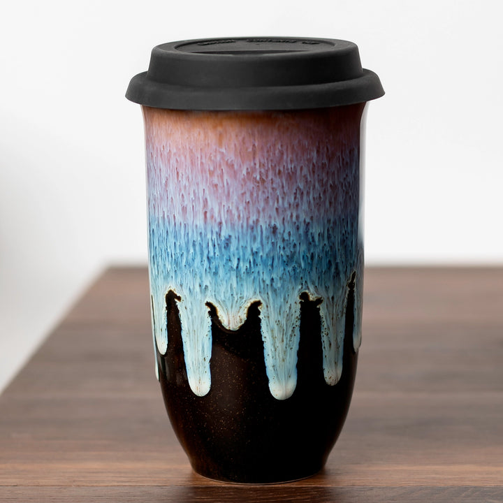 A ceramic travel tumbler with a black lid, featuring a dripping glaze design that transitions from dark brown at the bottom to light blue and dusty pink at the top, set on a wooden surface against a white background.