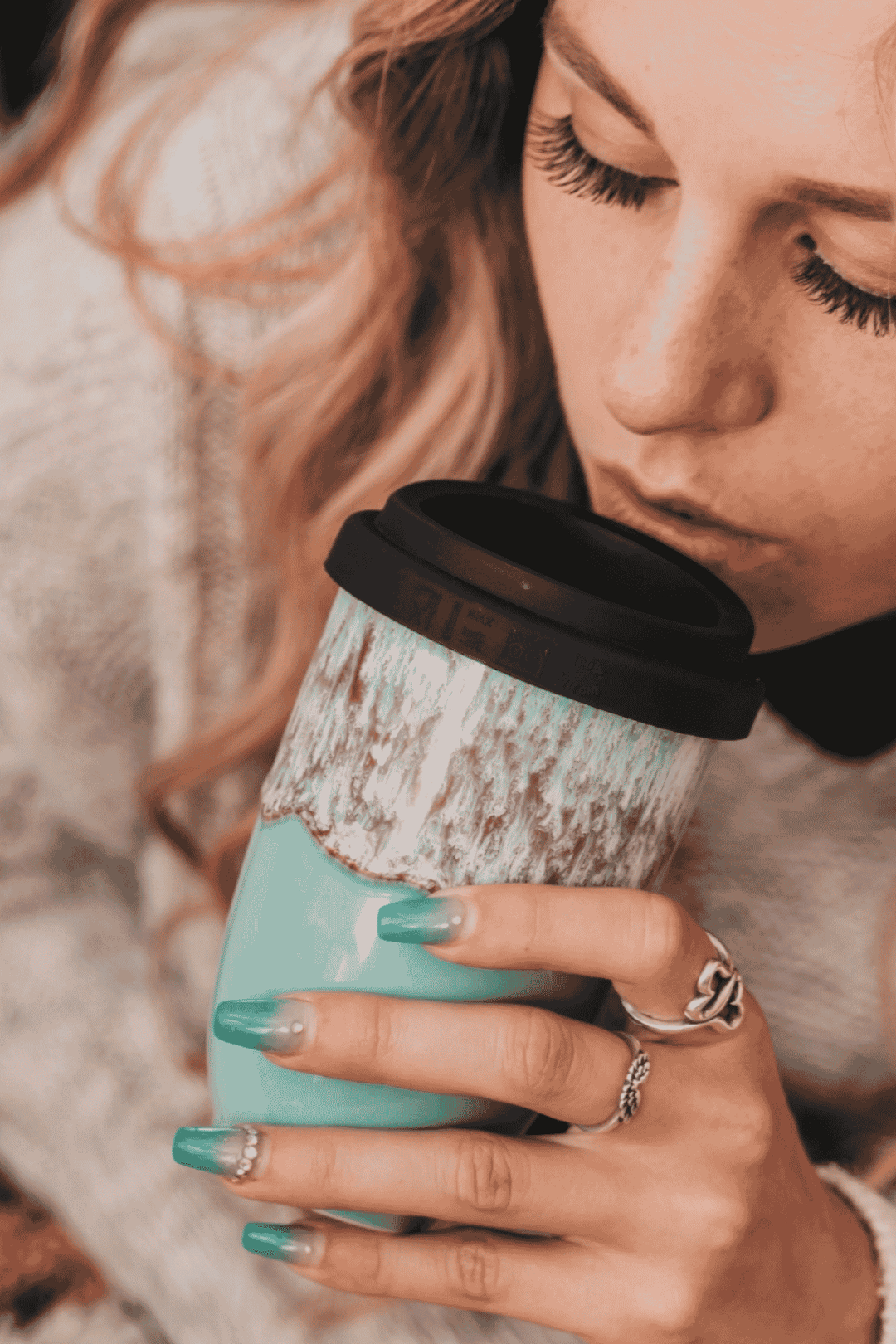 A close-up lifestyle shot of a woman holding a mint green travel tumbler with a black lid and a distressed white upper section, wearing silver rings and teal nail polish.