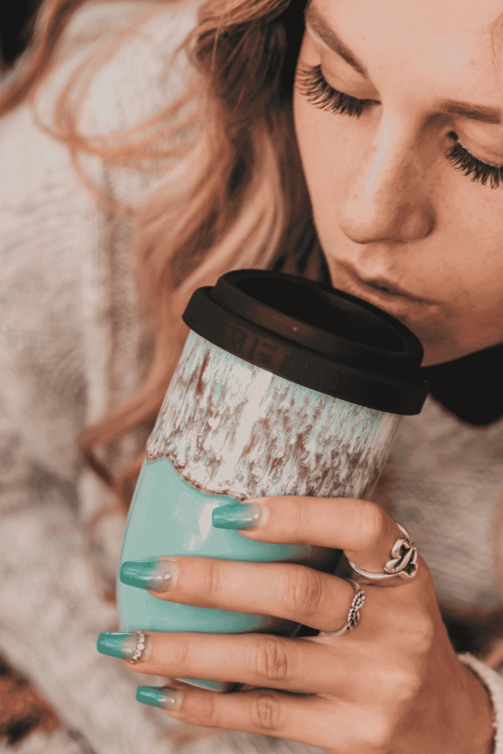 A close-up lifestyle shot of a woman holding a mint green travel tumbler with a black lid and a distressed white upper section, wearing silver rings and teal nail polish.