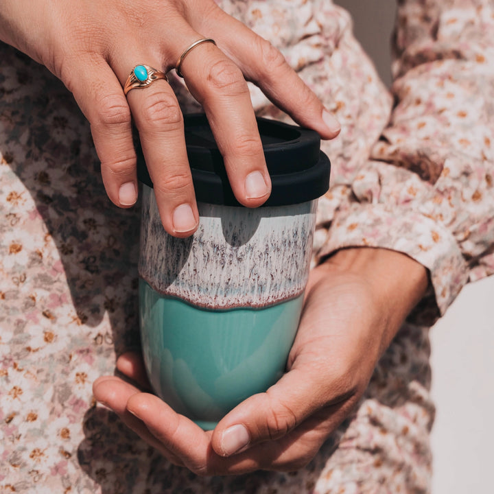 Person holding a teal mug with a black lid, wearing a floral-patterned garment.