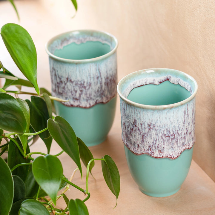 Two turquoise ceramic cups with a speckled design on a wooden surface, surrounded by green leaves.