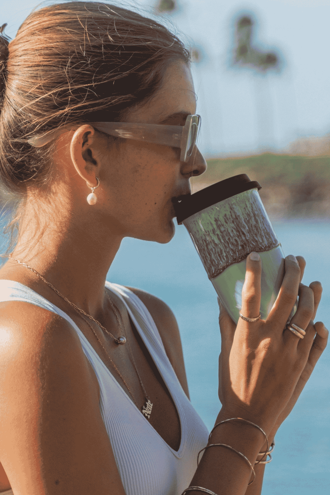 A woman in a white ribbed tank top and clear-framed sunglasses drinks from a reusable travel tumbler with a dark lid and a green-and-white marbled sleeve, set against a blurred outdoor background.