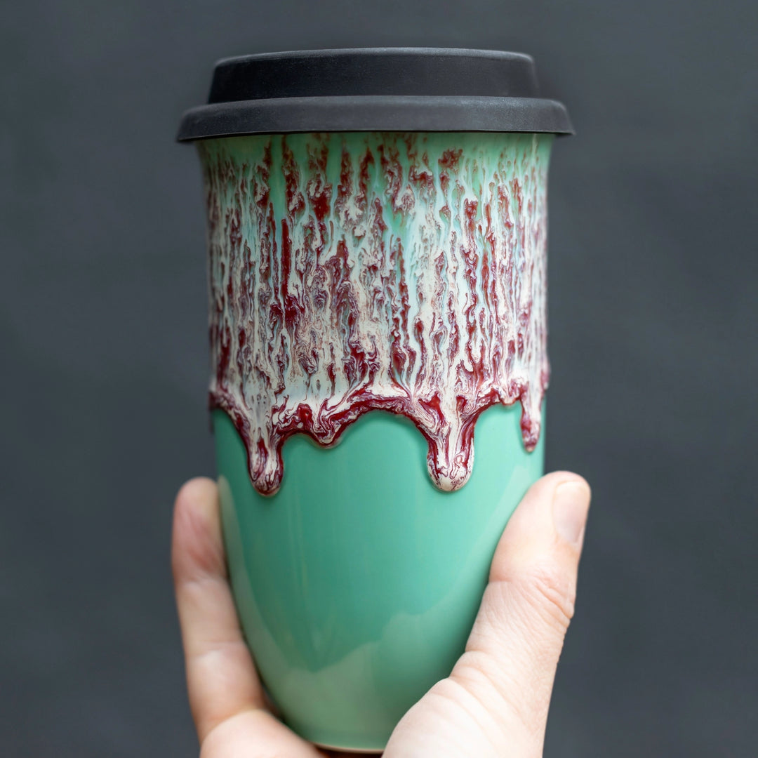 A hand holding a ceramic travel mug with a mint green body, a dripping red and white glaze pattern around the upper half, and a black lid, set against a dark gray background.