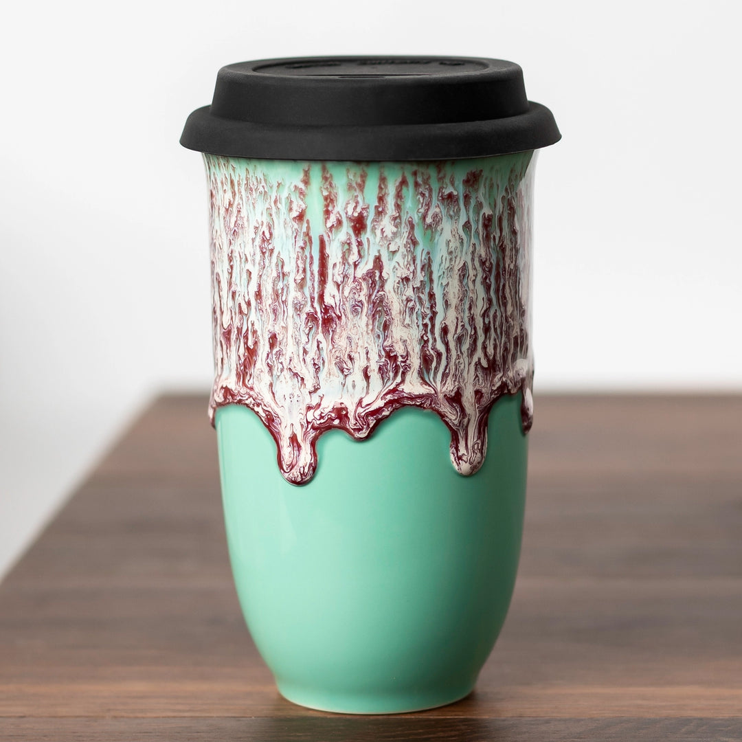 Turquoise ceramic tumbler with black lid and red and white abstract design on a wooden surface.