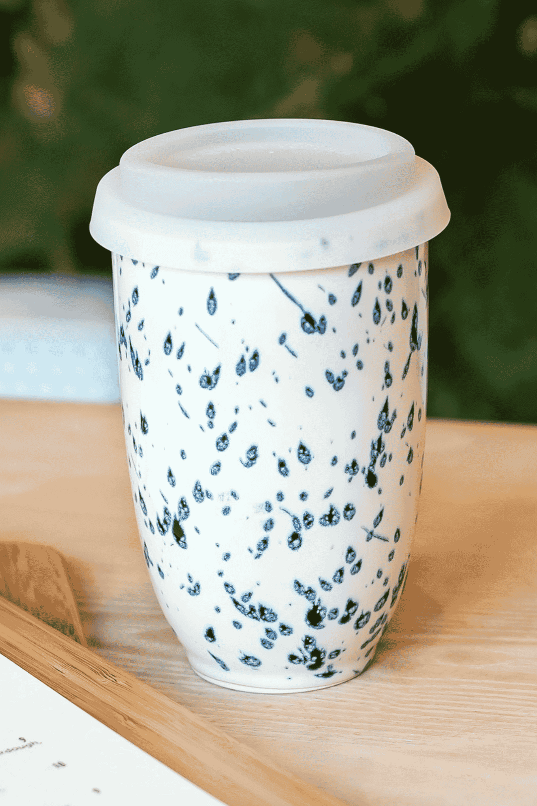 A reusable ceramic travel mug with a white lid, featuring a white body decorated with a black speckled pattern, resting on a light wooden surface.
