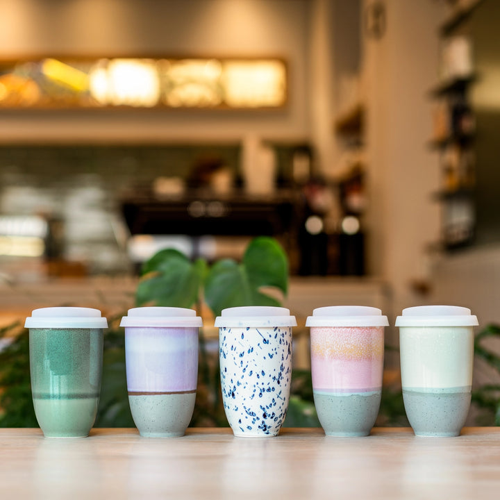 Five colorful ceramic cups with lids on a wooden surface in a blurred cafe setting.