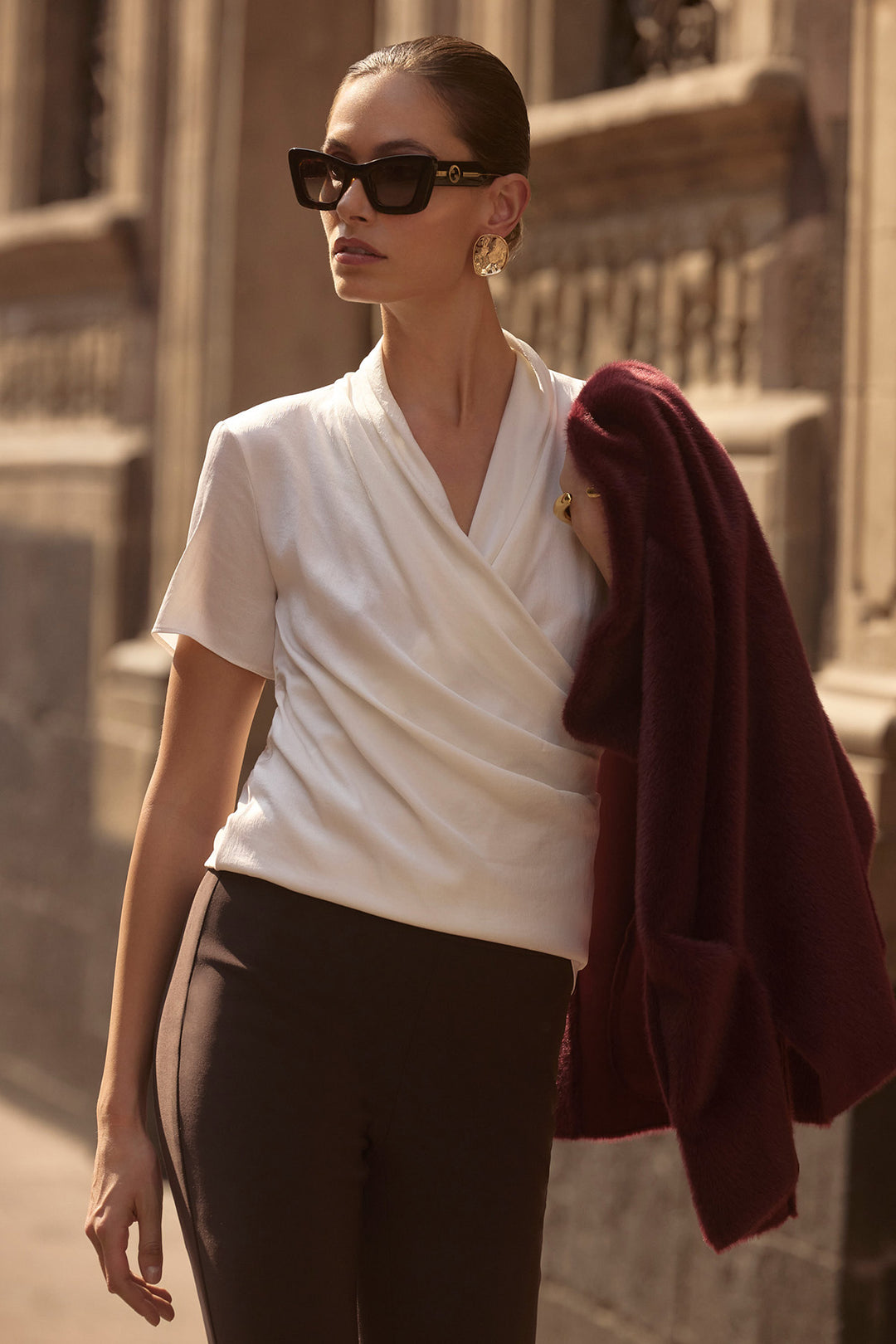 Woman wearing a white top and black pants with sunglasses, standing against a stone wall.