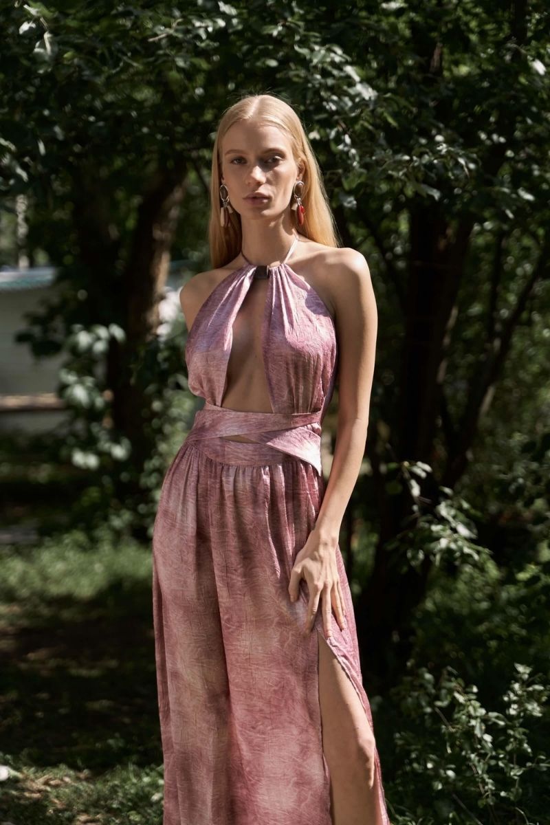 Woman wearing a pink dress with a halter neck and high slit, standing in a forested area.
