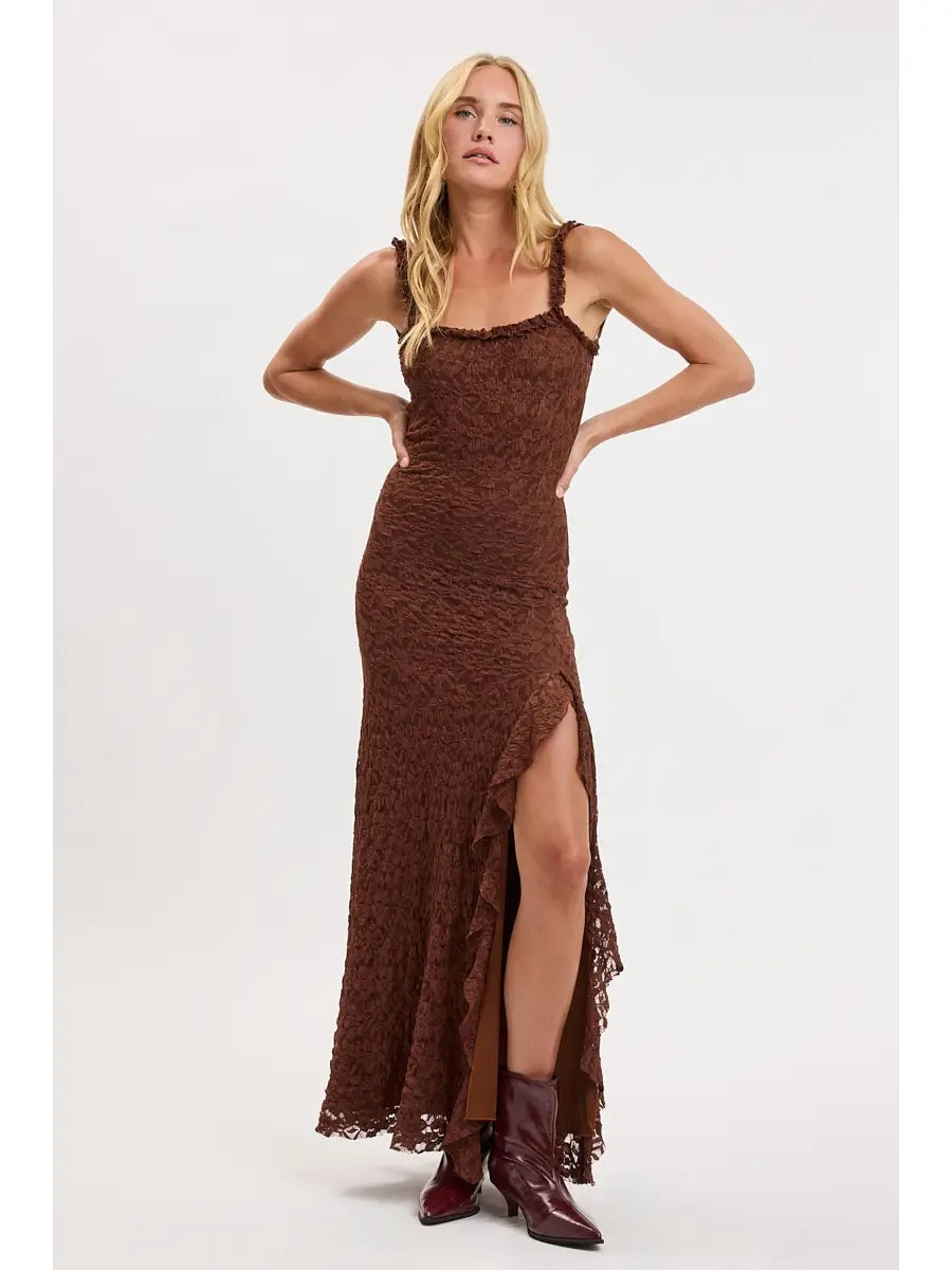 A blonde model wearing a brown lace maxi dress with a thigh-high side slit and dark brown pointed-toe boots, standing against a plain light gray studio background.