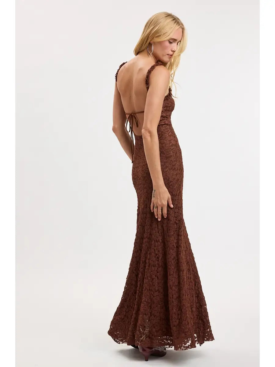A woman modeling a floor-length brown lace gown with a backless open back and tie detail, shown from the back against a plain light gray studio background.