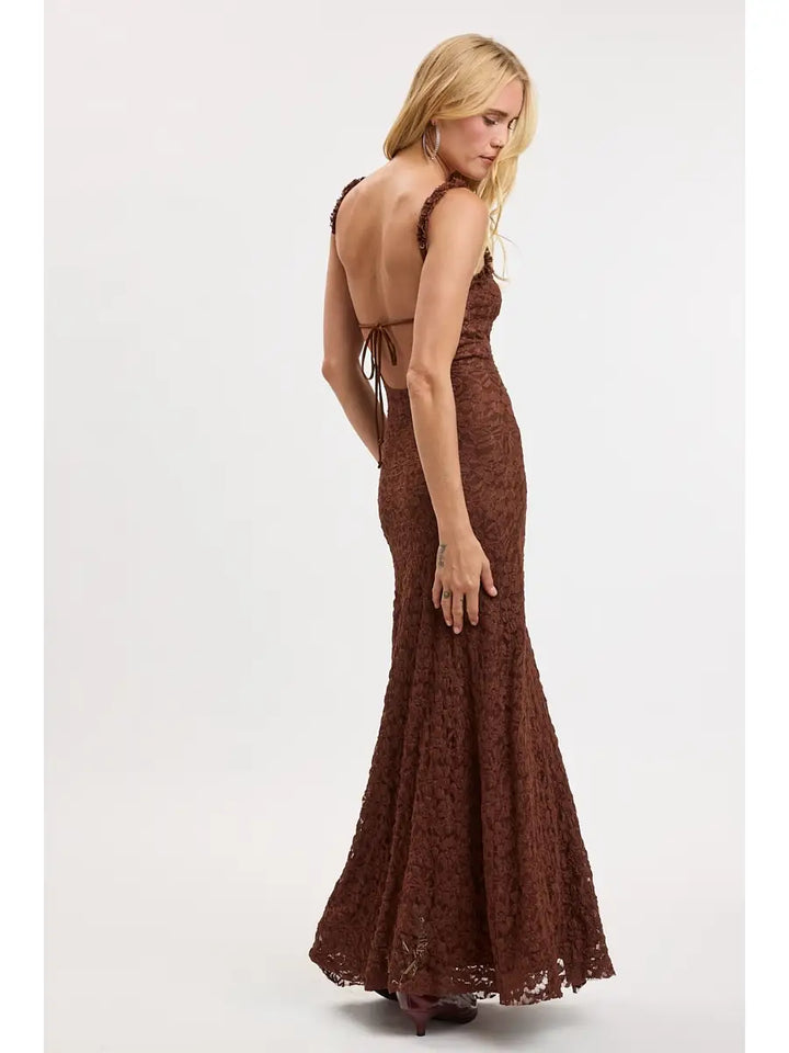 A woman modeling a floor-length brown lace gown with a backless open back and tie detail, shown from the back against a plain light gray studio background.