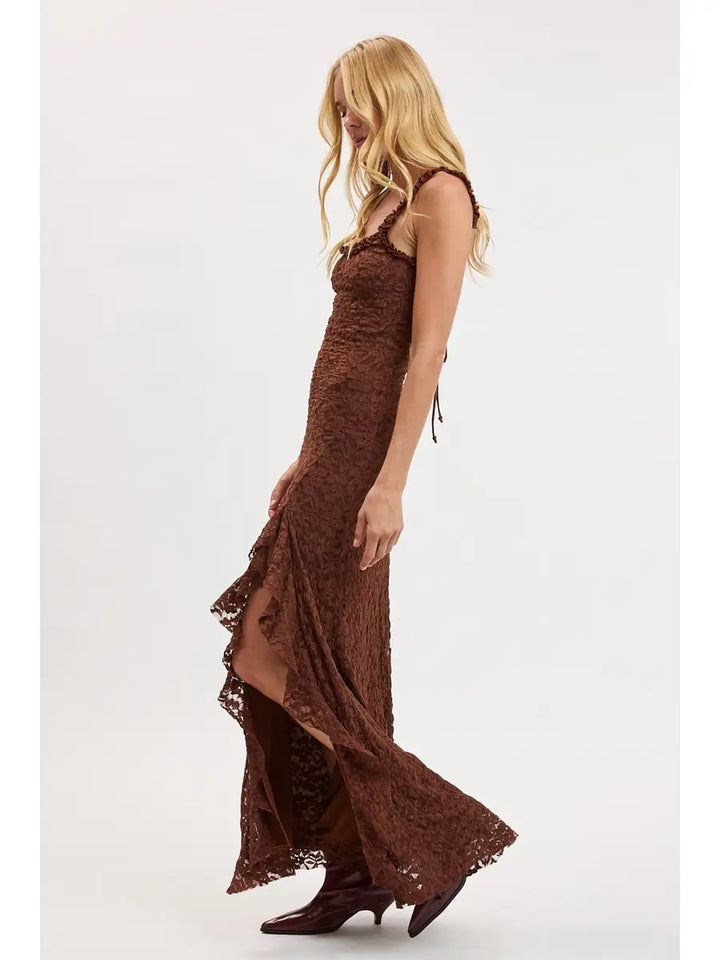 A blonde model wearing a brown lace maxi dress with side tie details and brown heeled boots, shown from a side profile against a plain white studio background.
