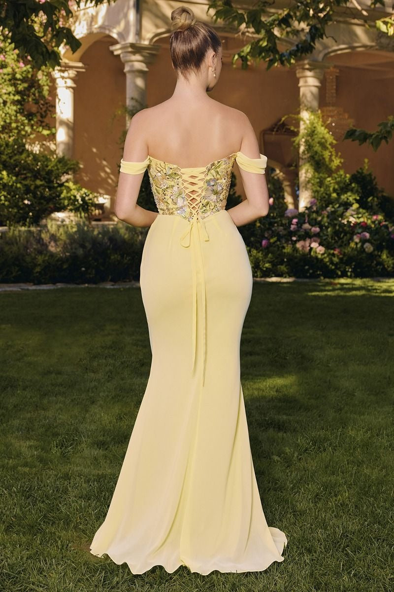 Woman in a yellow evening gown standing outdoors with greenery and a building in the background