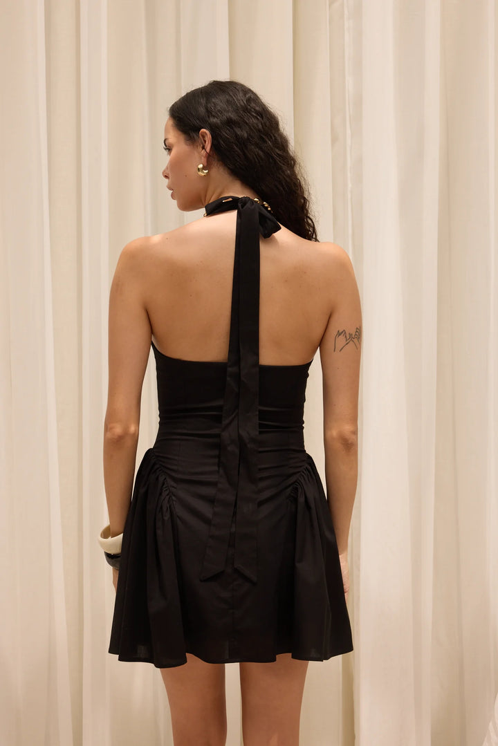 Woman wearing a black halter neck dress with a bow detail, standing against a beige curtain.