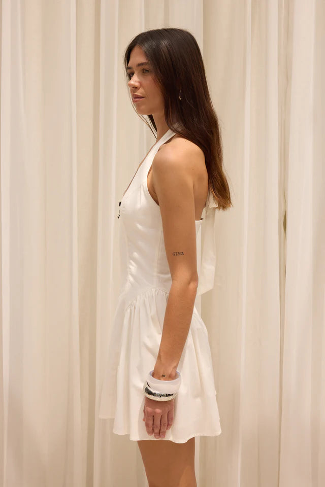 Woman wearing a white dress standing against a beige curtain.