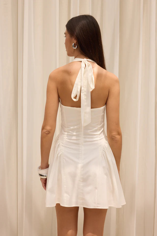 Woman wearing a white dress with a halter neck design against a beige curtain background