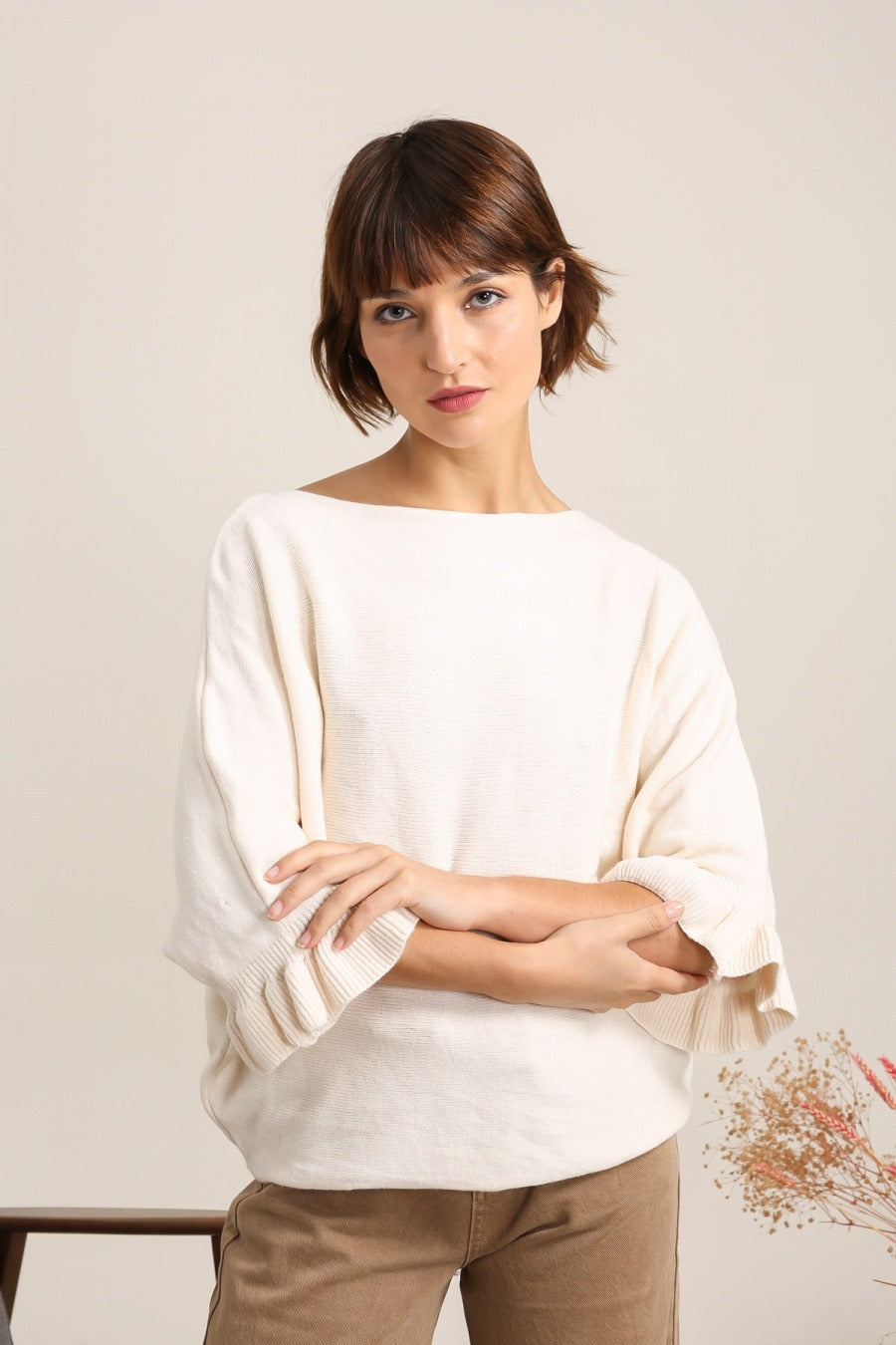 Woman wearing a white sweater and beige pants against a plain background