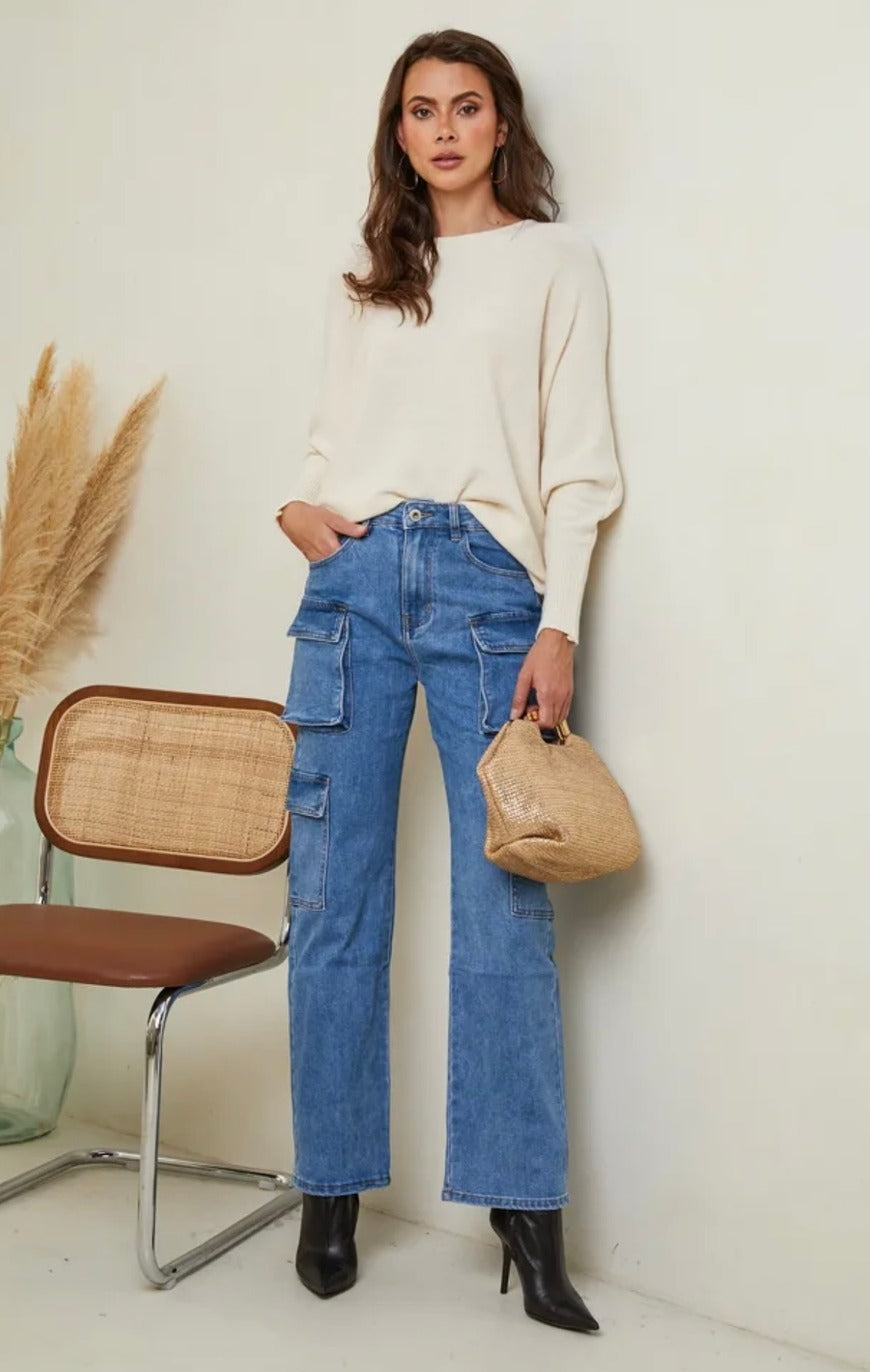 Woman wearing a white sweater and blue jeans, holding a beige handbag, standing in a minimalistic room.