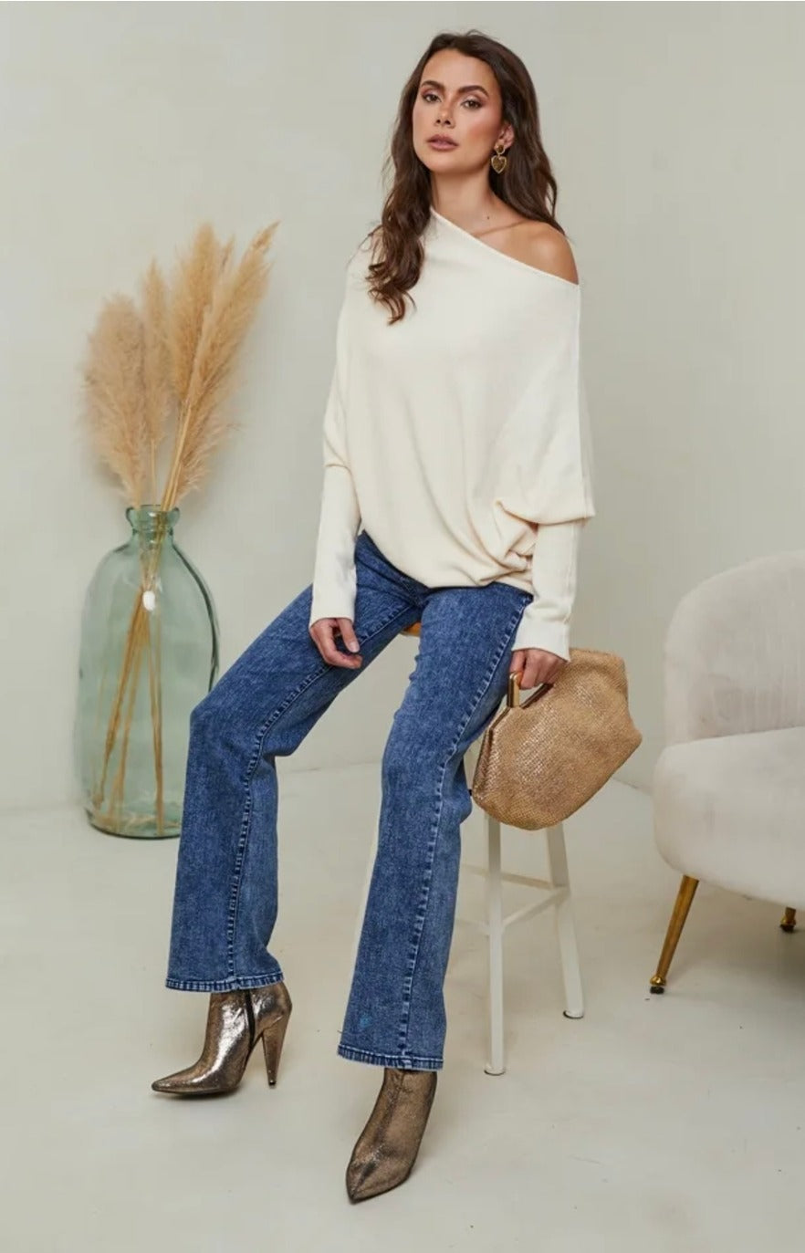 Woman wearing a white off-shoulder top and blue jeans sitting on a stool with a neutral background.