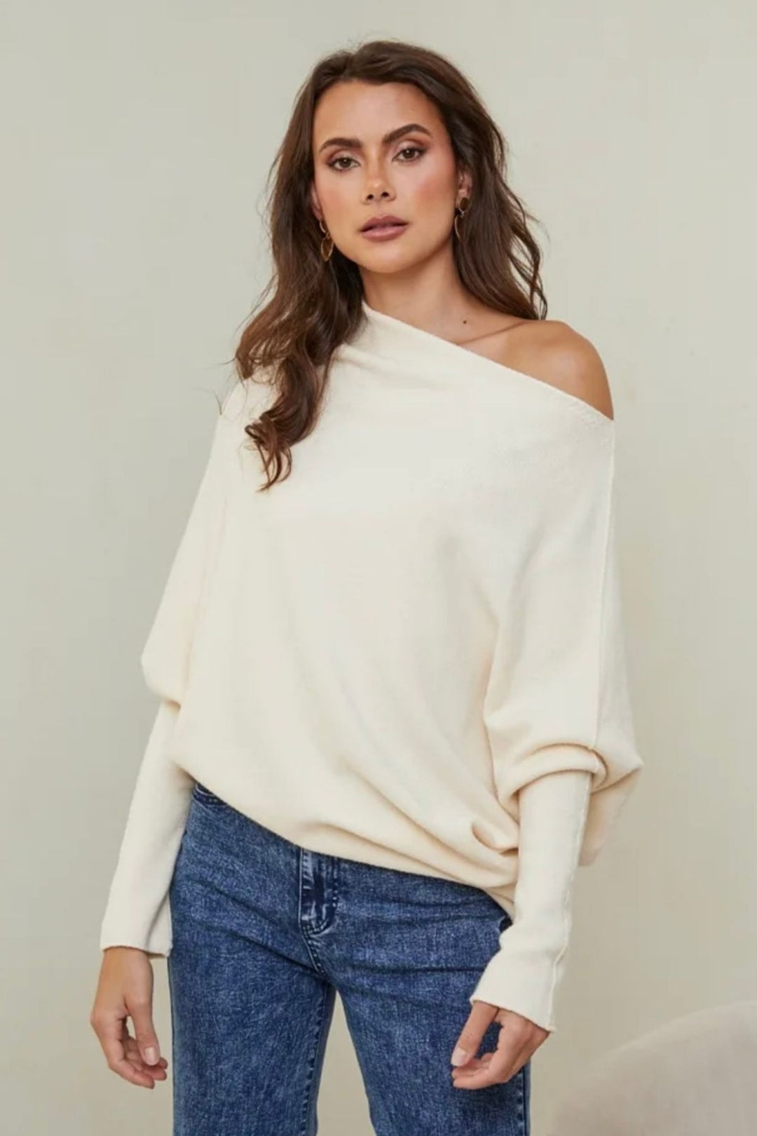Woman wearing a white off-shoulder sweater and blue jeans against a plain background