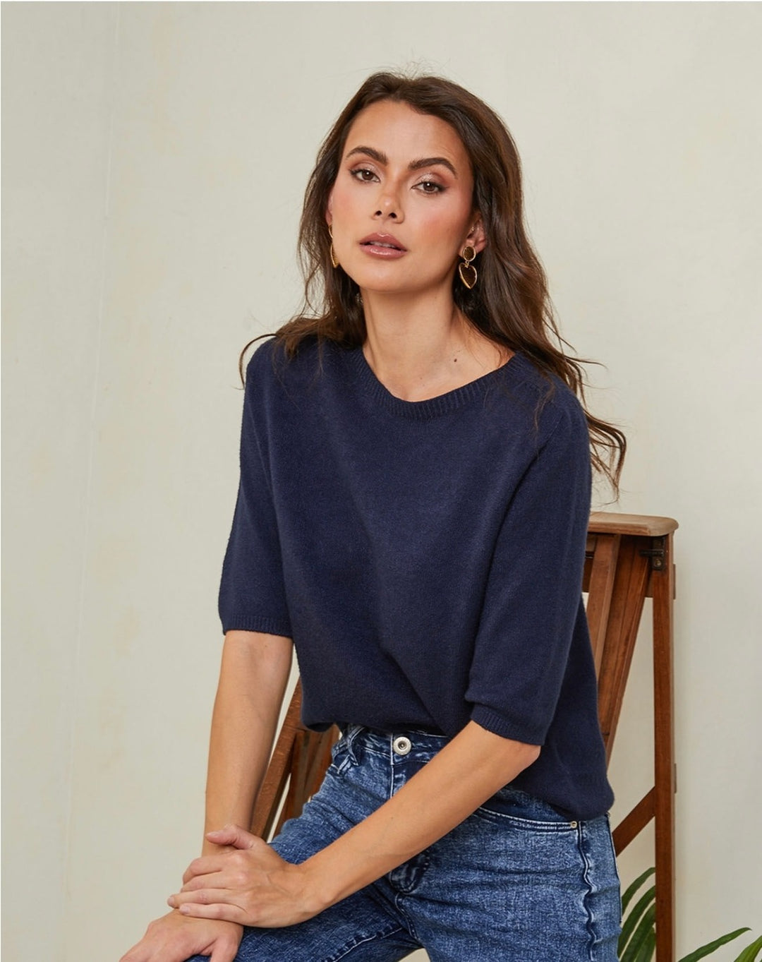 Woman wearing a navy blue sweater and jeans sitting on a wooden chair against a beige wall.
