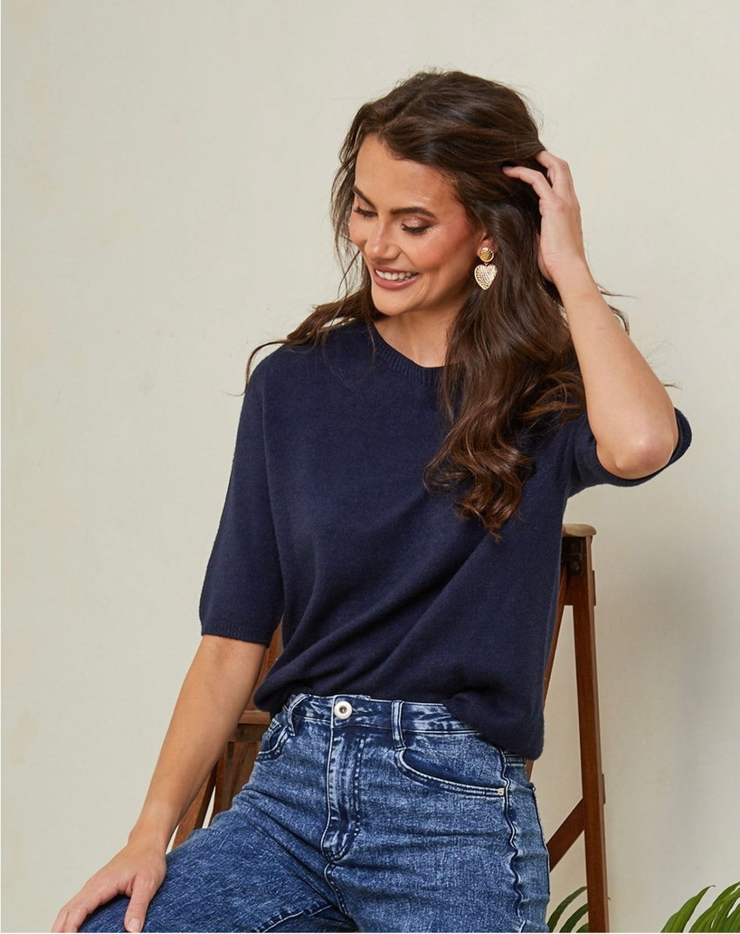 Woman wearing a navy blue top and jeans sitting on a chair against a neutral background