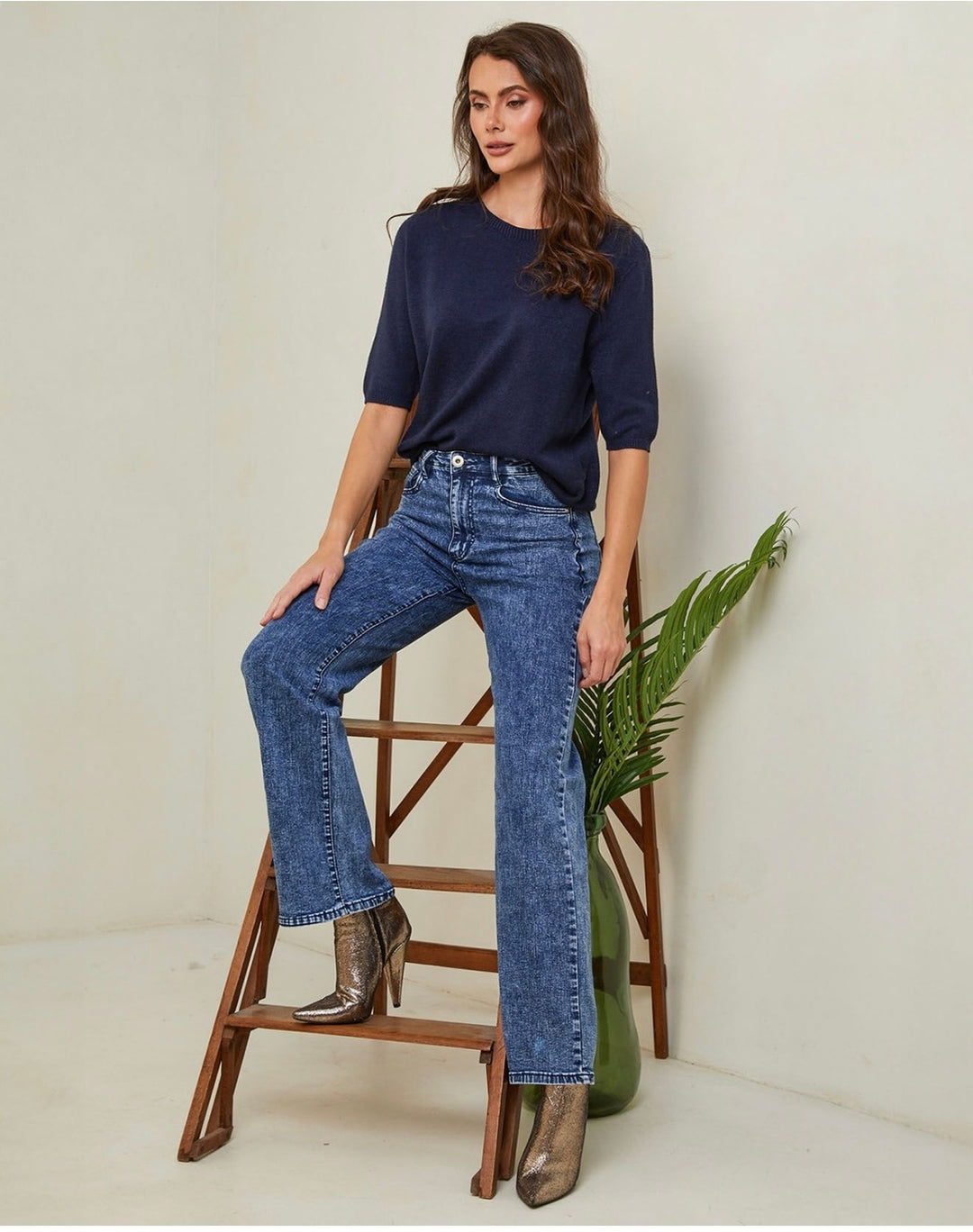 Woman wearing a navy top and blue jeans sitting on a wooden stool with a plant in the background.