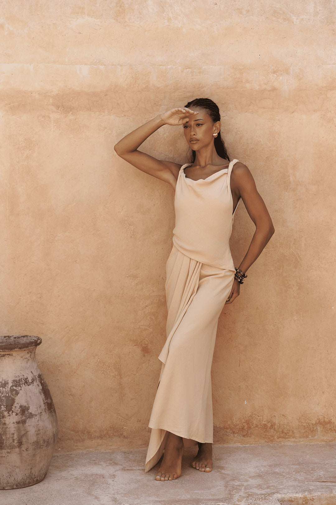 Woman in a beige dress standing against a textured wall with a rustic pot.