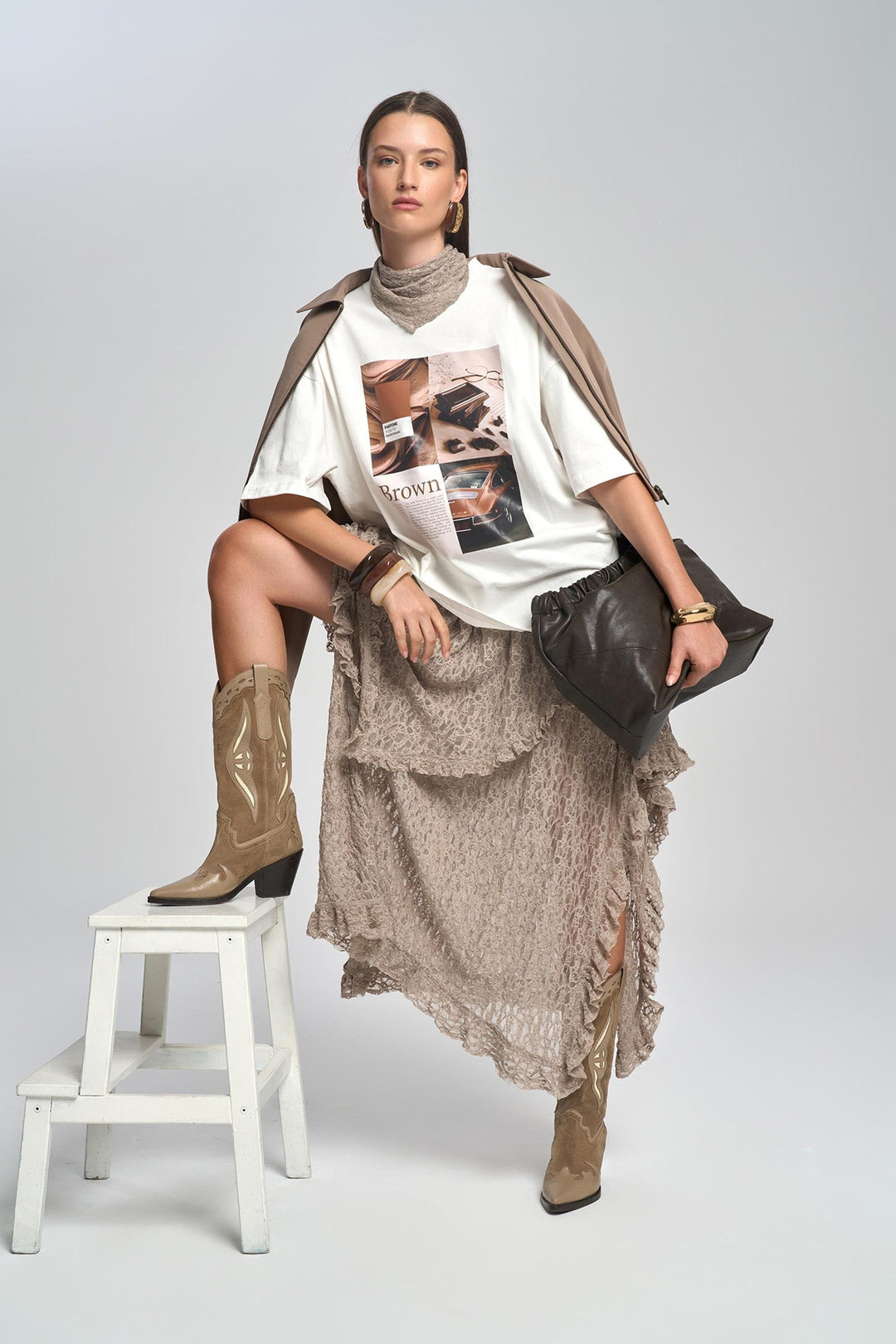 Woman sitting on a stool wearing a white t-shirt with graphics, beige skirt, and brown boots on a plain background.