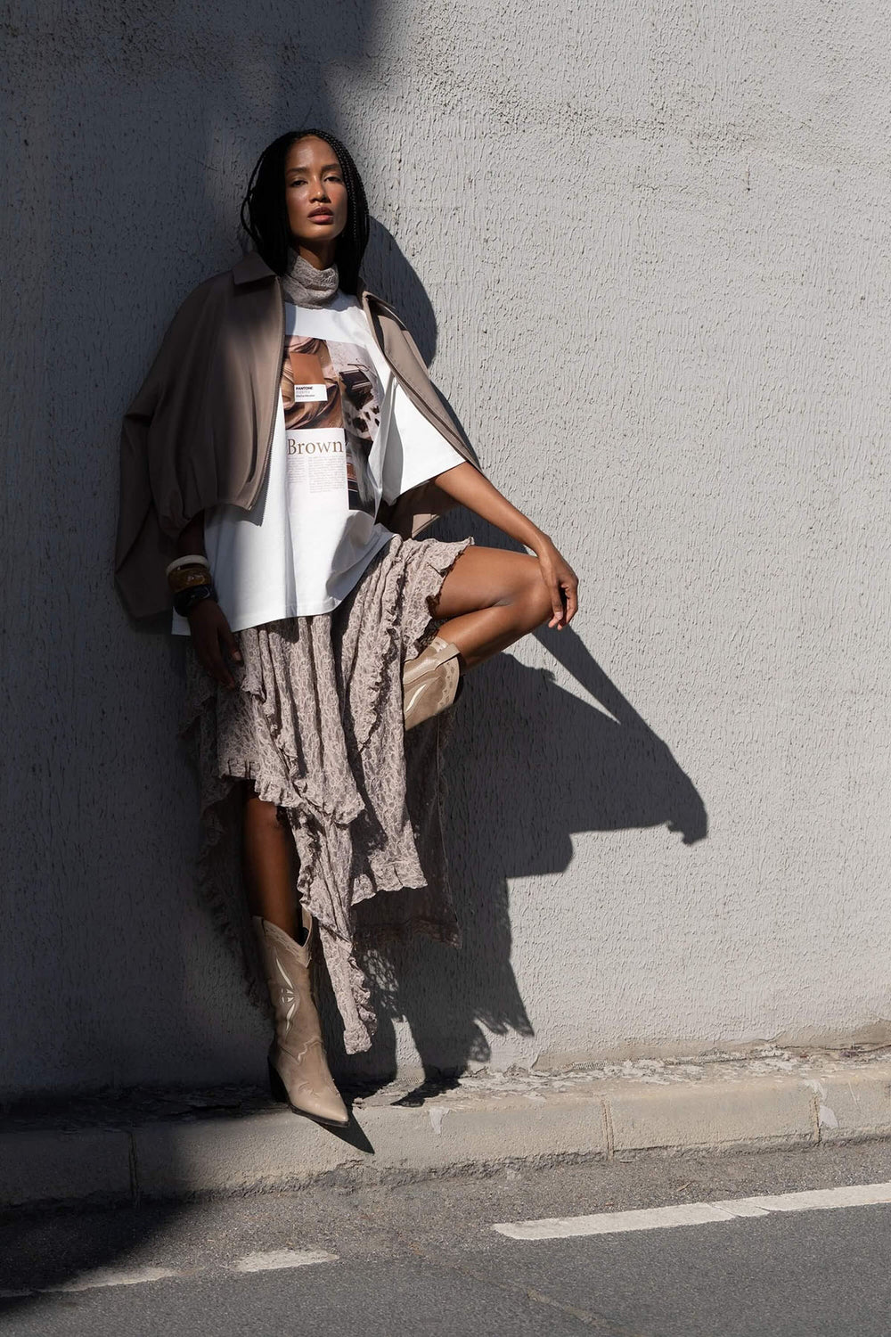 Woman in a casual outfit with a graphic t-shirt, beige cardigan, and boots against a gray wall.