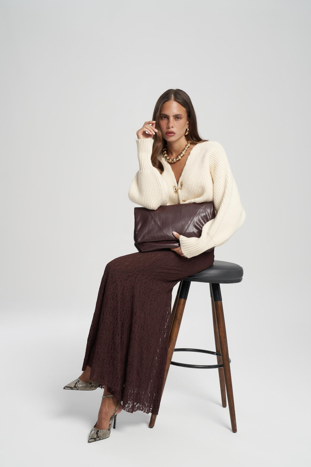 Woman sitting on a stool wearing a white sweater and brown dress against a white background