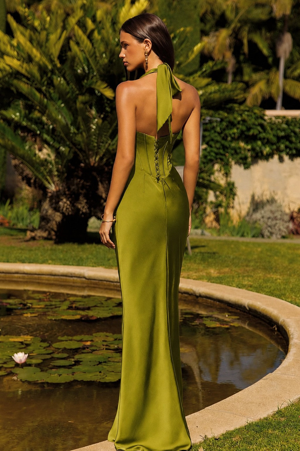 A woman stands outdoors by a pond wearing a floor-length olive green evening gown with a tie-back neckline and open back, photographed in a garden setting.