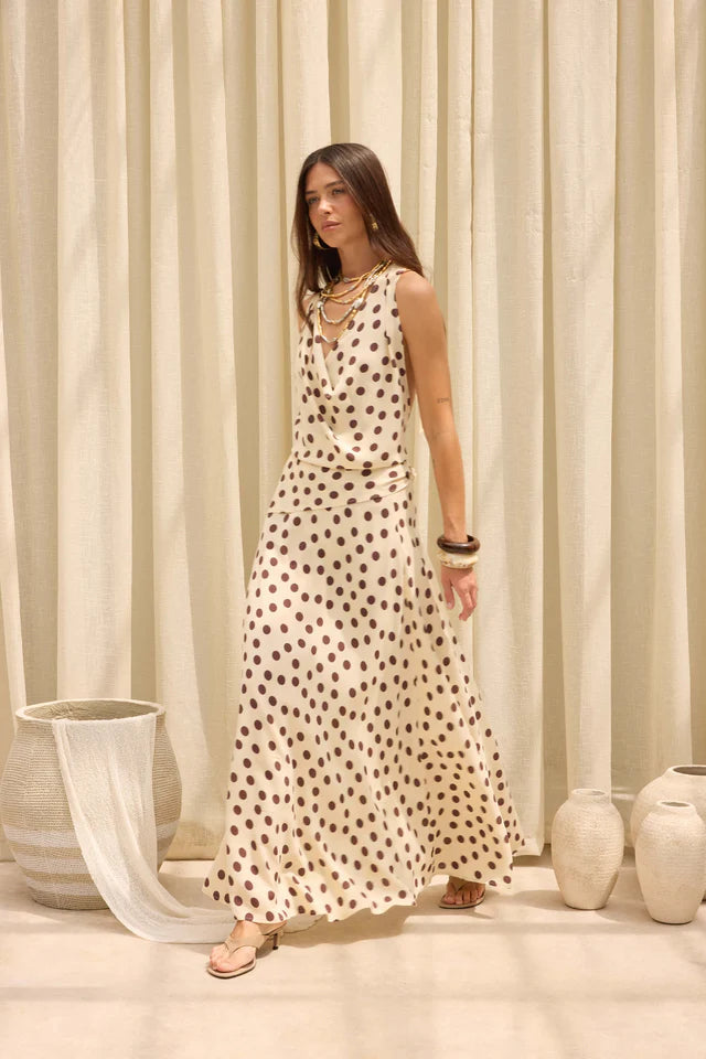 A woman modeling a sleeveless, floor-length cream maxi dress with a dark brown polka dot pattern, standing in front of a draped beige curtain backdrop with decorative ceramic vases.