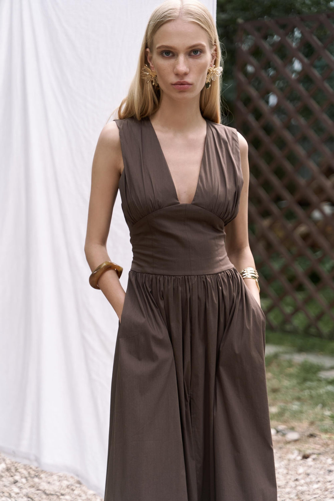 Woman wearing a brown dress standing outdoors with a white curtain and wooden lattice in the background.