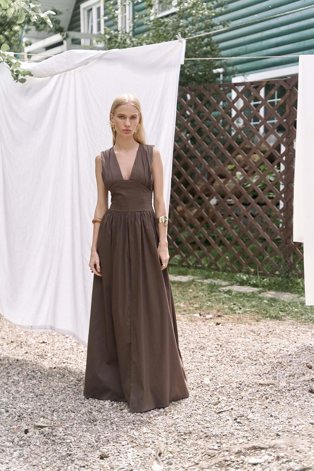 Woman in a long brown dress standing outdoors with a white sheet and wooden lattice in the background.