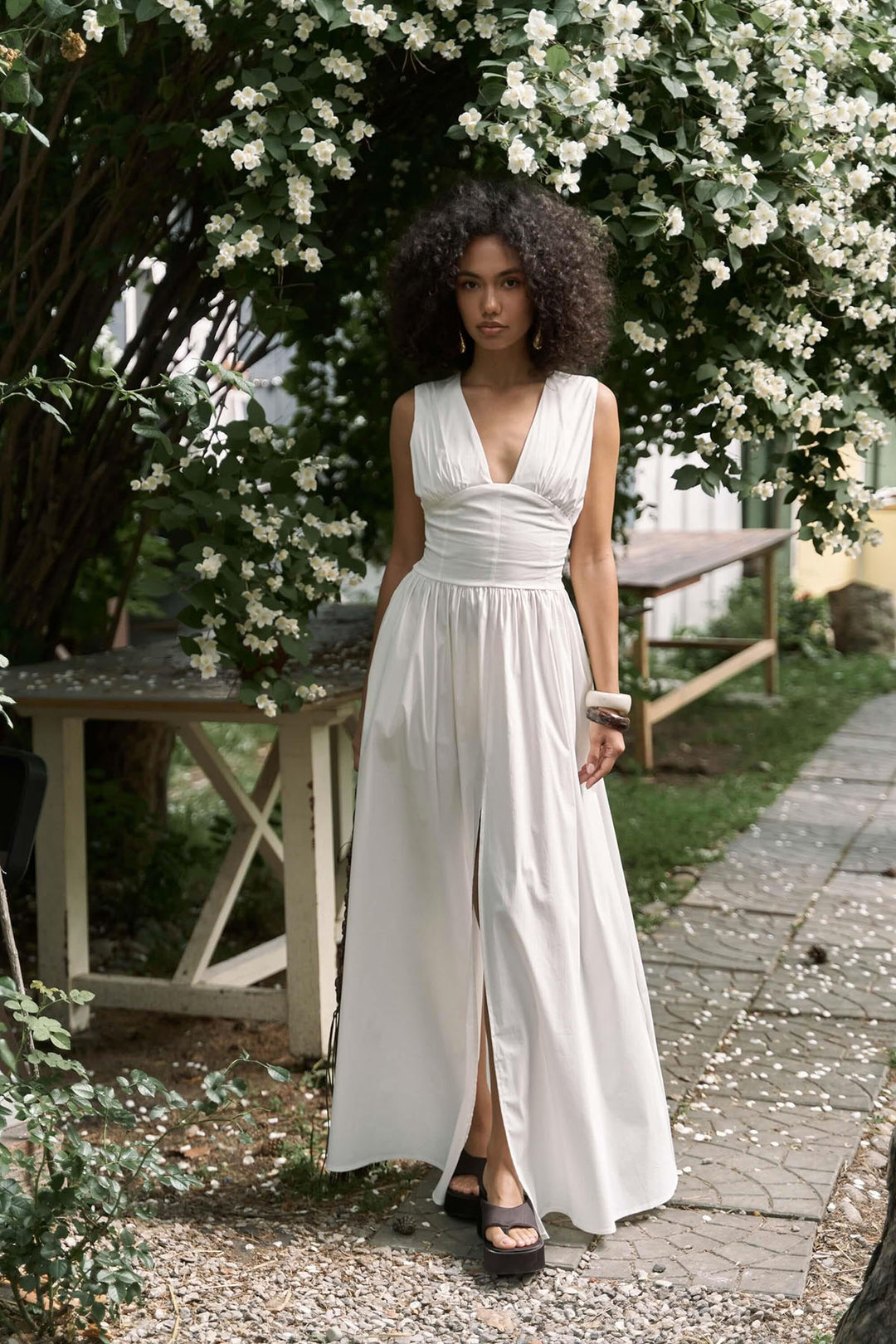 Woman in a white dress standing in a garden with white flowers and a wooden table.