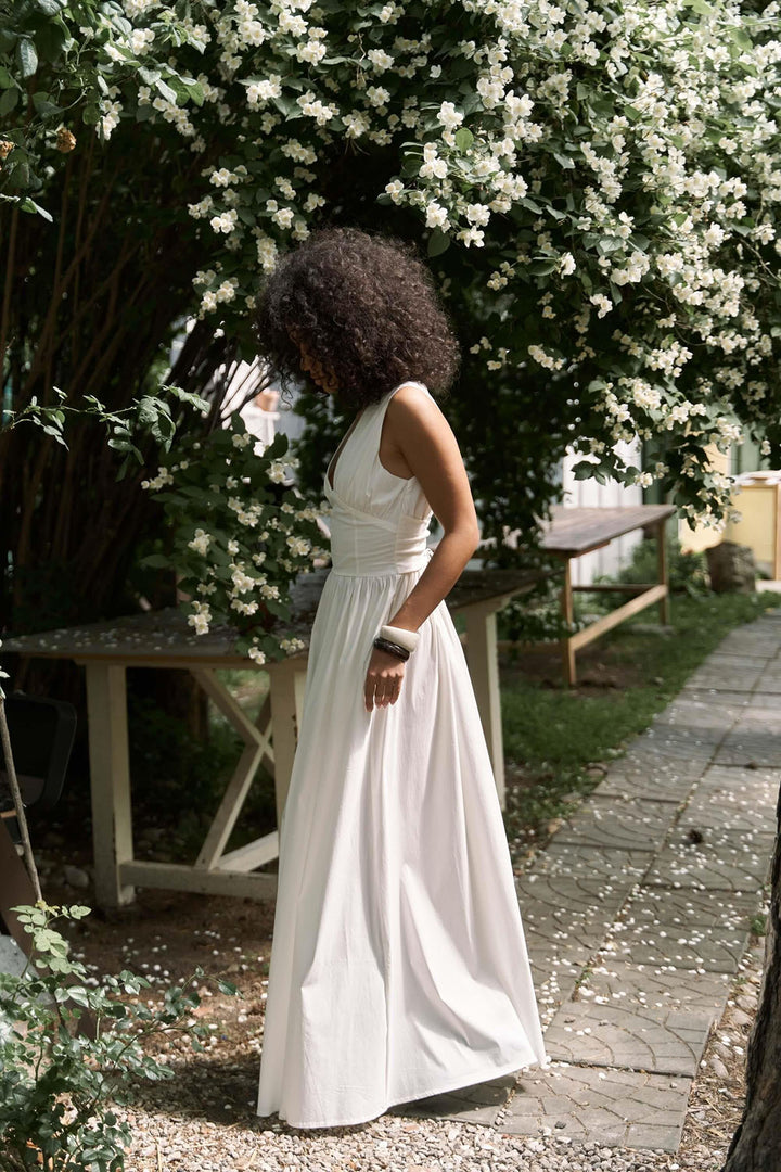 Woman in a white dress standing in a garden with white flowers and wooden furniture.