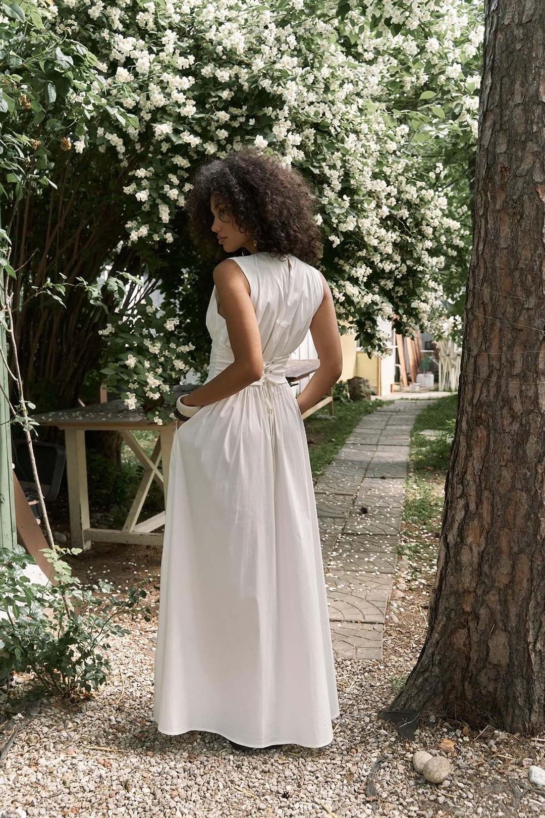 Woman in a white dress standing in a garden with white flowers and a tree.
